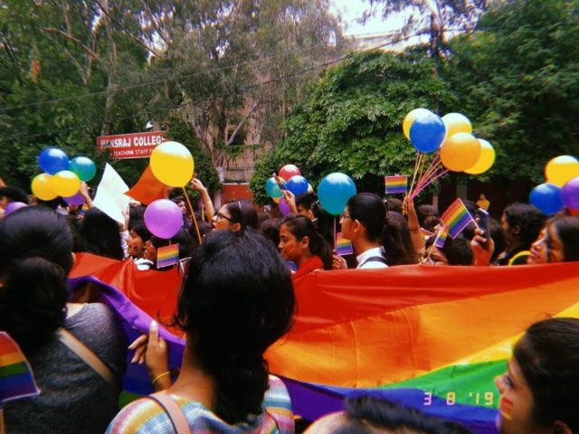 A pride parade passing underneath a sign saying, "Hansraj Hostel"